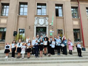 Grupa skolēnu dažādos vecumos stāv uz pakāpieniem pie brūnas ēkas, katrs turot sertifikātu. Virs ēkas durvīm redzams uzraksts: "DIĀNA 3. PAMATSKOLA", un uz durvīm ir logo "TSP".