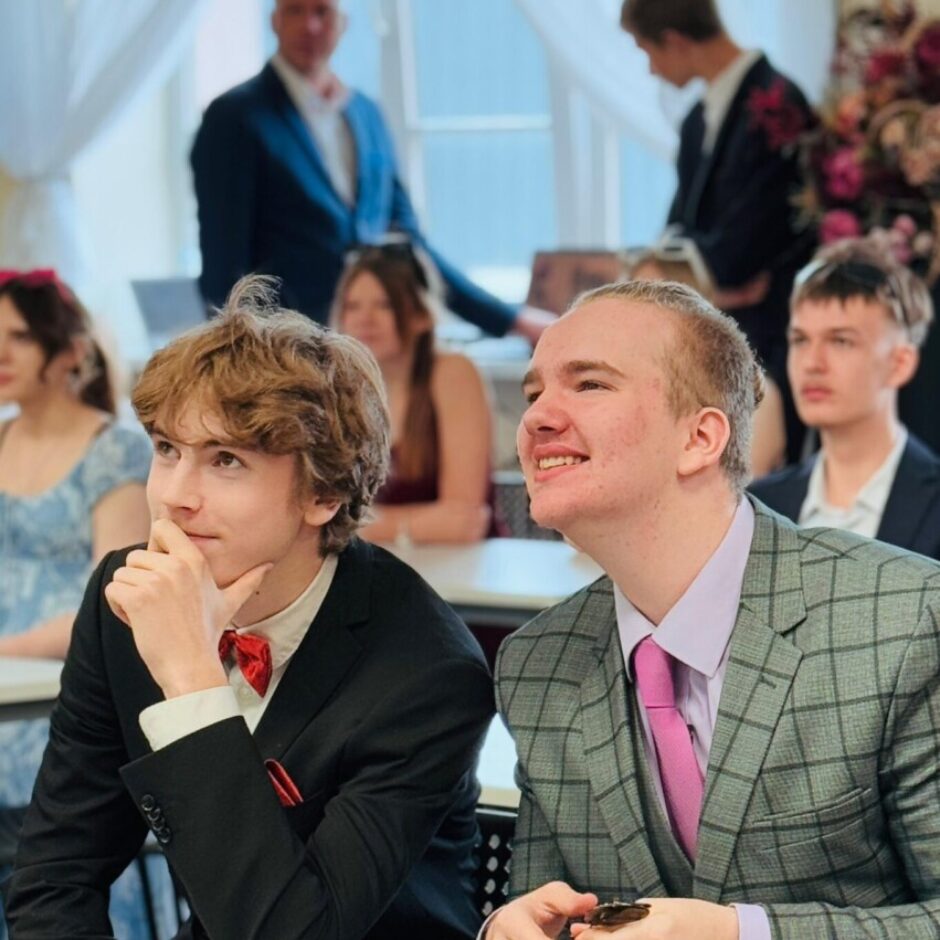 Divi jauni vīrieši formālā apģērbā sēž pie galdiem pasākumā. Kreisais puisis melnā uzvalkā ar sarkanu tauriņu domīgi skatās uz augšu. Labais puisis pelēkā rūtainā žaketē ar rozā kaklasaiti smaida, skatīdamies uz augšu, un rokās tur mazu tumšu priekšmetu. Fonā redzami citi cilvēki.