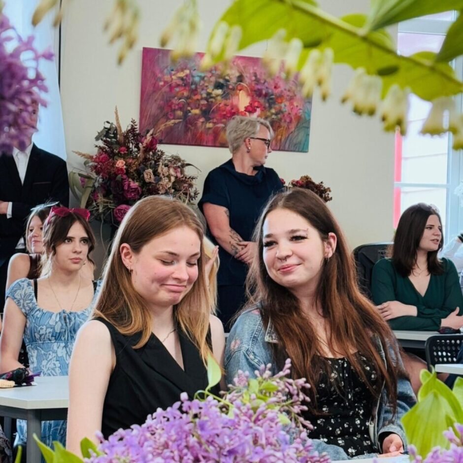 Divas jaunas sievietes sēž pasākuma zālē, priekšplānā purpura ziedu pušķis. Viena no sievietēm smaidot skatās kamerā, otra skatās uz leju ar vieglu smaidu. Fonā redzami citi cilvēki un abstrakta glezna.