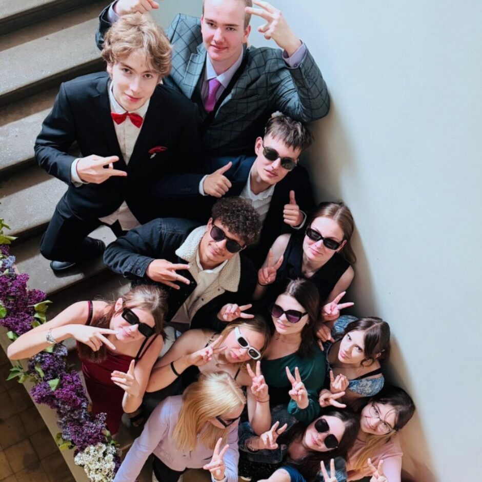 Liela jauniešu grupa pozē uz kāpnēm, skatoties uz augšu kamerā. Vairums nēsā saulesbrilles un rāda miera zīmes vai īkšķus uz augšu. Daži ģērbušies formāli, citi brīvāk. Kāpņu margas rotātas ar violetiem un baltiem ceriņu ziediem.
