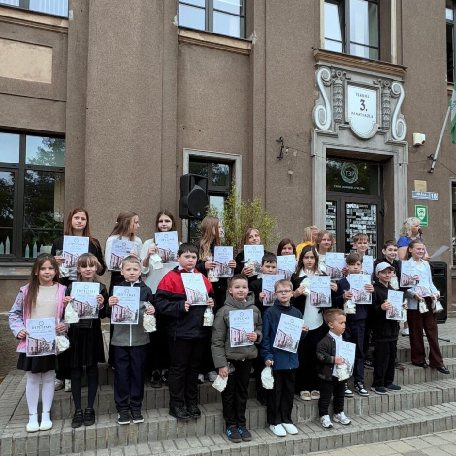 Skolēnu grupa, turot diplomus un mazus baltus maisiņus, stāv uz kāpnēm pie ēkas, uz kuras redzams uzraksts 