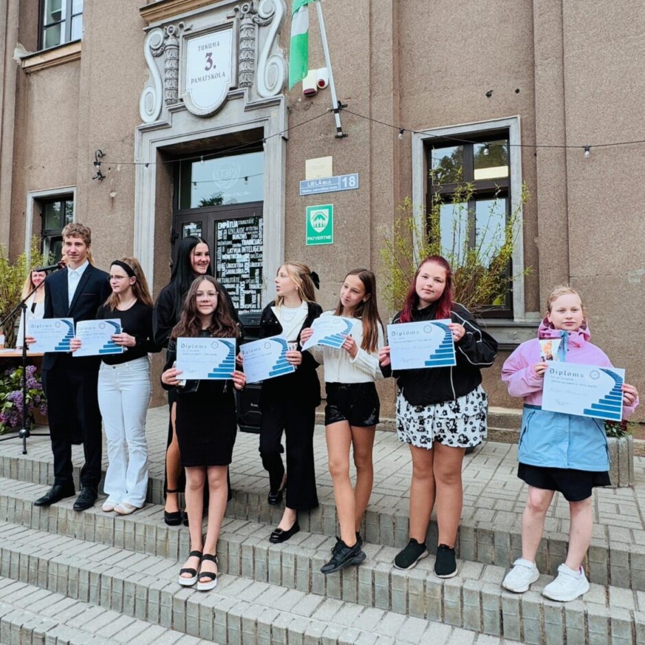 Grupa deviņu jauniešu, tostarp viens puisis un astoņas meitenes, stāv uz akmens kāpnēm Tukuma 3. pamatskolas priekšā. Visi tur rokās diplomus, uz kuriem redzams vārds 