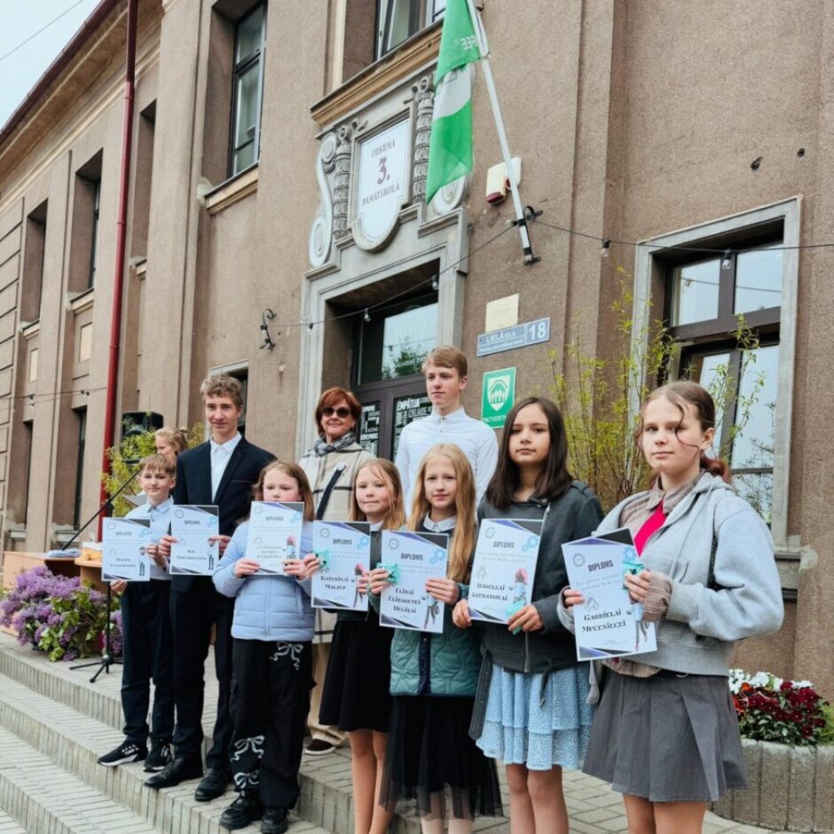 Bērnu un pieaugušo grupa stāv uz kāpnēm pie skolas Tukuma 3. pamatskolas ēkas, turot rokās diplomus.