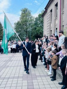 Jauns vīrietis uzvalkā nes tirkīzzilu karogu ar uzrakstu "ECO-" un "EEE", soļojot garām skolas ēkai garām stāvošiem dažāda vecuma skolēniem, blakus sievietei. Skolēni vēro gājienu.