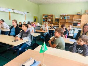 Vairāki bērni un pieaugušie sēž gaiši zaļā klasē pie koka galdiem, uzmanīgi raugoties uz priekšu. Aizmugurē redzami plaukti ar grāmatām un zīmējumi pie sienas. Priekšplānā uz galda ir zaļš karodziņš ar baltu logo un uzrakstu "FEE".