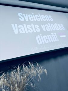Ekrānā projicēts teksts: "Sveiciens Valsts valodas dienā!" ar sirds formas Latvijas karogu blakus, un zemāk: "Valsts valoda – mūsu kopīgā telpa." Ekrāna apakšējā daļā priekšplānā redzams kaltētu graudaugu kūlis.