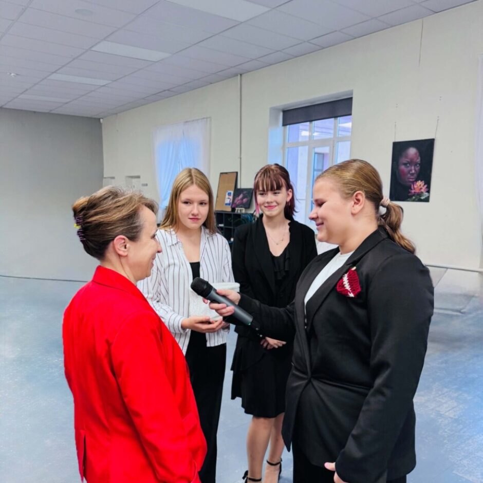 Četras sievietes telpās: jauna sieviete melnā žaketē ar Latvijas karoga piespraudi intervē sievieti sarkanā žaketē ar mikrofonu. Divas citas jaunas sievietes vēro sarunu.